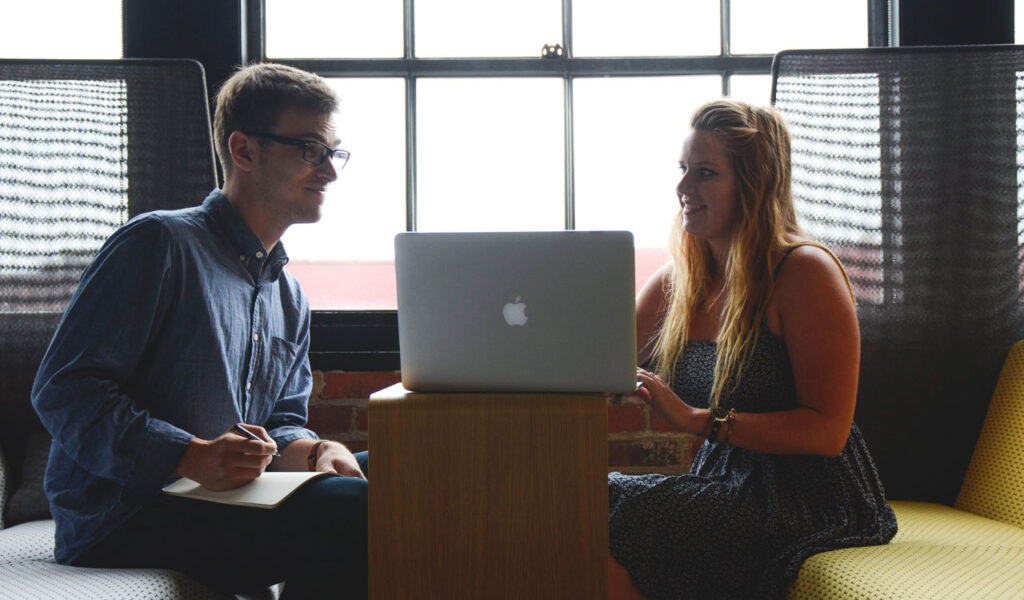 Two people in casual clothes sitting at a table with a laptop, discussing a project. Two people in casual clothes sitting at a table with a laptop, discussing a project.